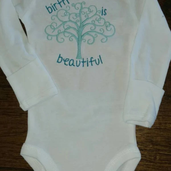 Tree Of Life Machine Embroidery stitch-out: White baby onesie with green tree and text design on a wooden surface