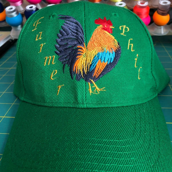 Golden Rooster Embroidery Design stitch-out: Green cap with embroidered rooster design and text on a crafting table.